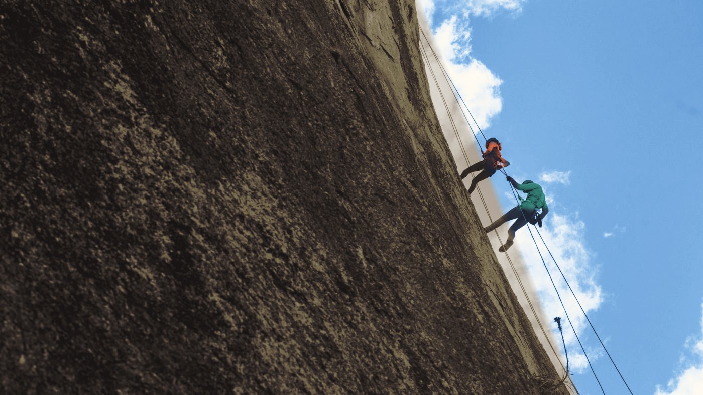 Two people on ropes against a steep rock face, working their way down together