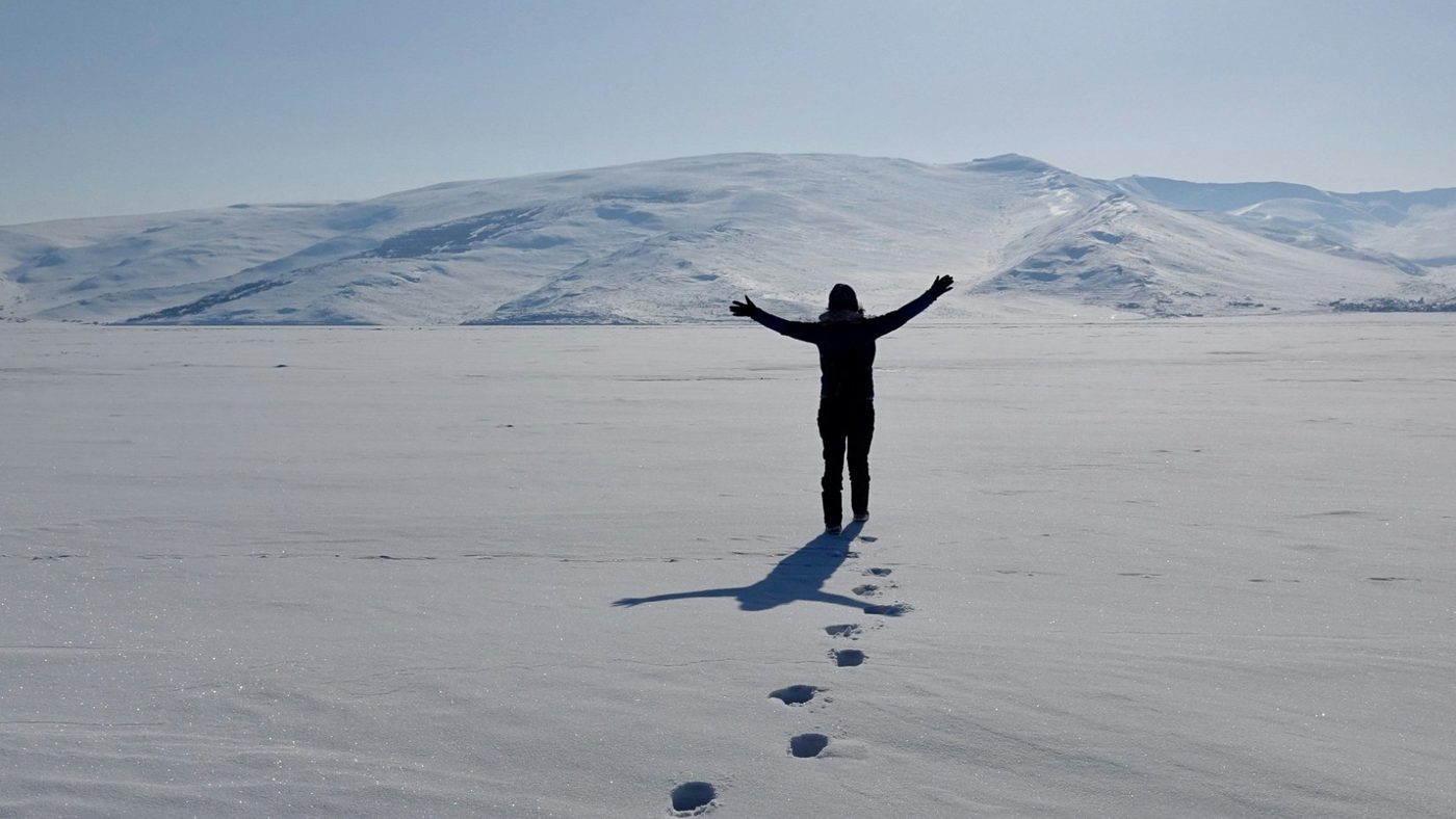 A person with arms open standing on a wide snowfield, with a line of footprints leading to them and snowy hills behind